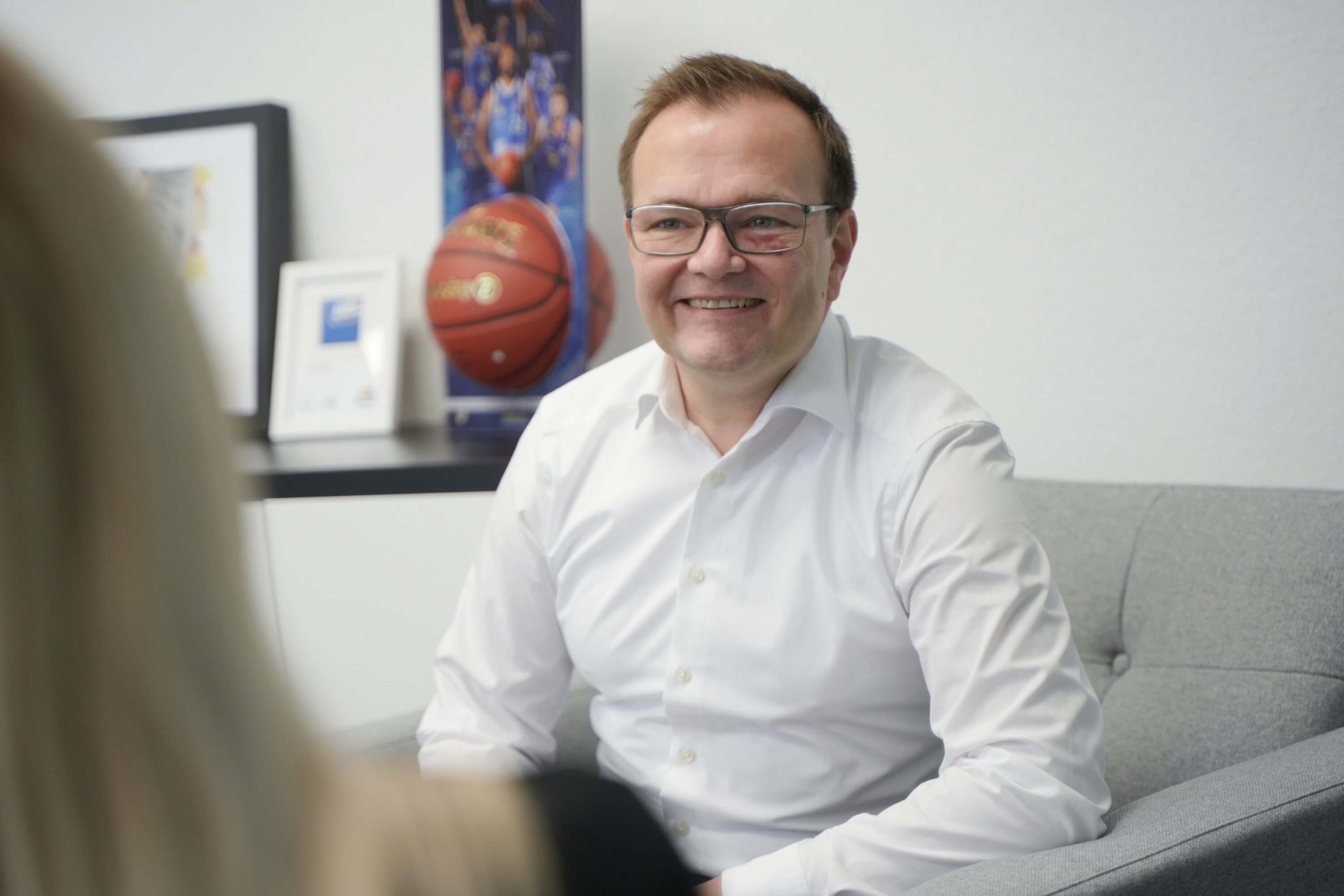 CEO Michael Zimmermann von Zimmermann IT-Solutions, ein lächelnder Mann mit Brille in einem weißen Hemd, sitzt auf einem grauen Sofa während eines Vorstellungsgesprächs mit einer Bewerberin im schwarzen Blazer. Im Hintergrund sind ein Basketball und eine Urkunde sichtbar.