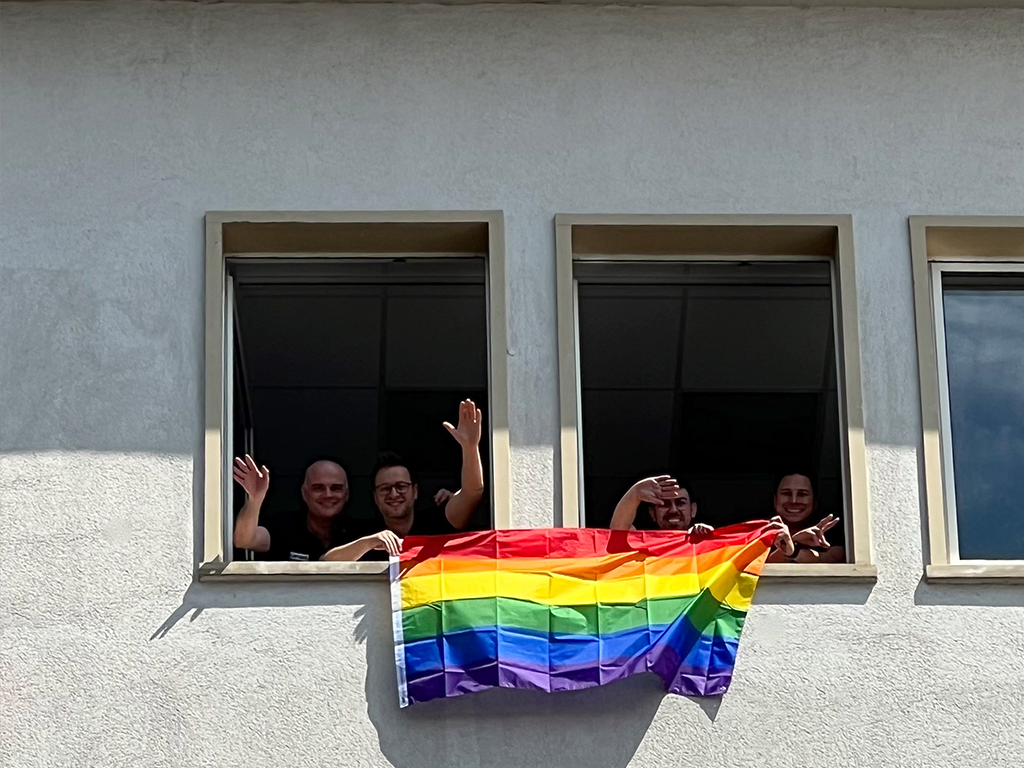 Vier Mitarbeiter halten eine farbenfrohe Pride-Flagge aus dem Fenster und genießen einen fröhlichen Moment zusammen.