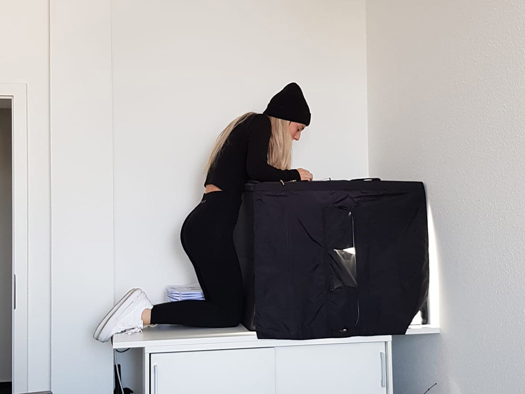 Vanessa macht Produktfotos an einer schwarzen Fotobox. Sie trägt eine schwarze Mütze und weiße Schuhe und kniet auf einem Tisch, um von oben in die Box zu schauen.