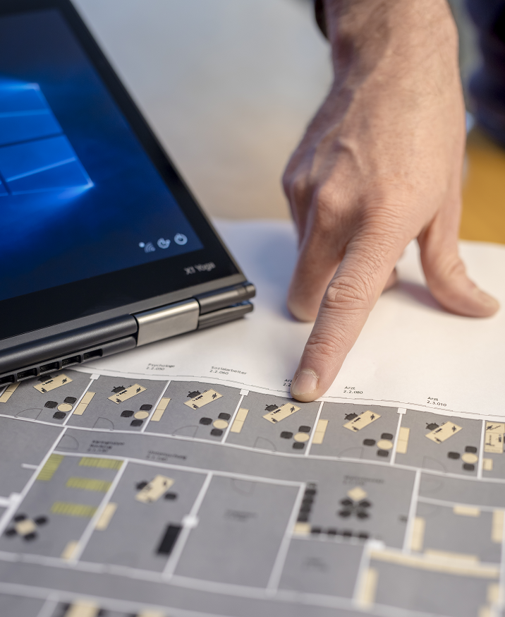Bild von einer männlichen Hand, welche auf einen Architechtenplan zeigt. Nebendran liegt ein Lenovo Yoga, welches für einen Planungsakt stehen soll.