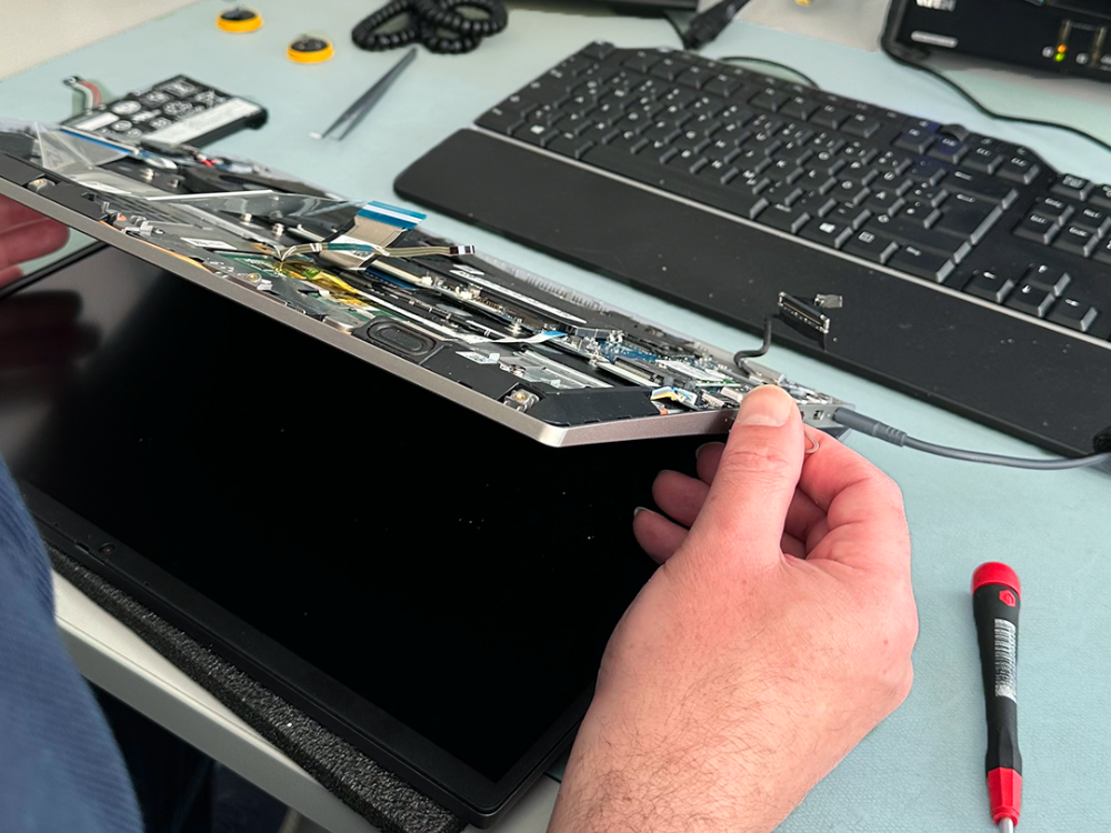 In der Abteilung Technik wird ein Notebook repariert. Der Mitarbeiter hat bereits die Rückseite abgeschraubt und hält es über die blaue Technik-Sicherheitsmatte.